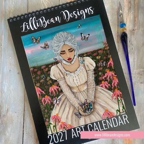 A 2027 art calendar by LilliBean Designs, featuring a colorful illustration of a woman in a cream dress surrounded by pink flowers and butterflies on the cover, displayed on a rustic wooden surface next to a paintbrush.
