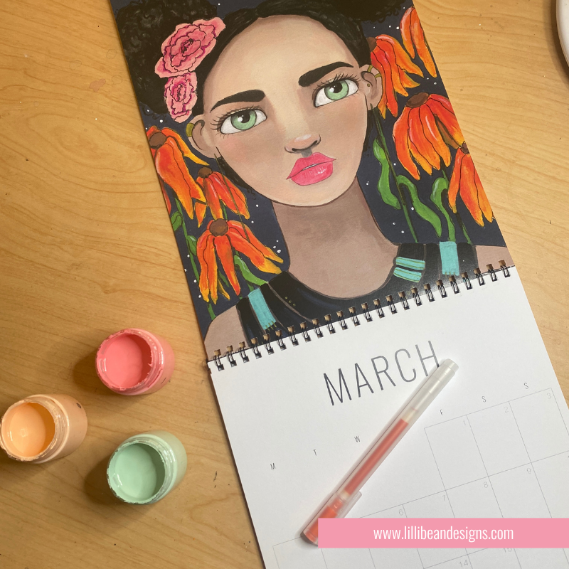 A spiral-bound March calendar lies open on a wooden table, featuring a colorful painted portrait of a woman with flowers in her hair. Paintbrushes and small paint pots are placed beside it, and a pink website watermark appears at the bottom.