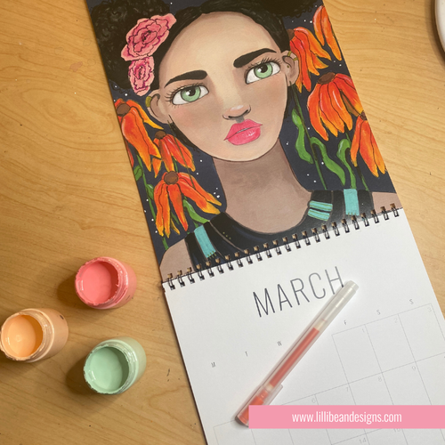A spiral-bound March calendar lies open on a wooden table, featuring a colorful painted portrait of a woman with flowers in her hair. Paintbrushes and small paint pots are placed beside it, and a pink website watermark appears at the bottom.
