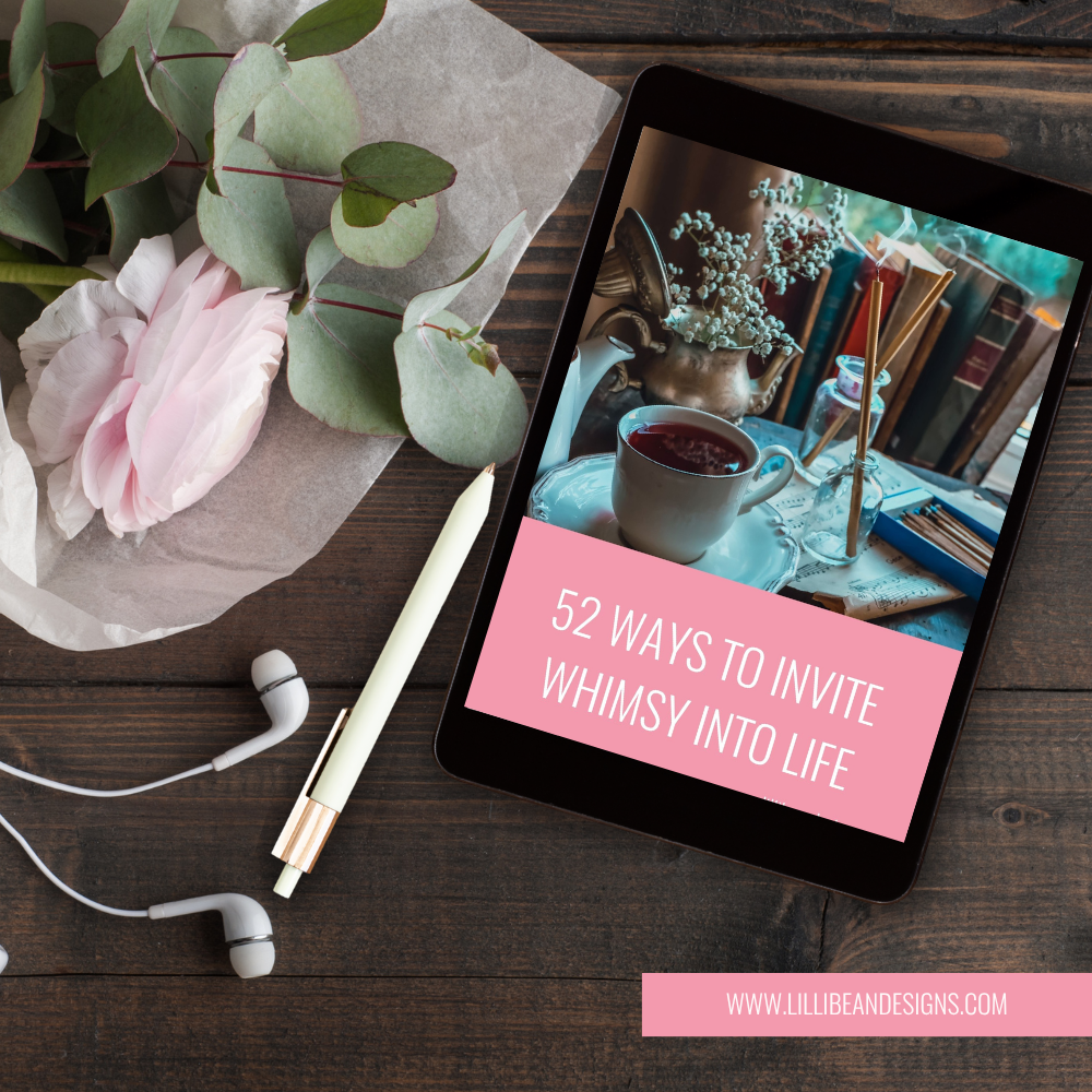 A tablet on a dark wooden desk displaying a pink graphic with the text '52 WAYS TO INVITE WHIMSY INTO LIFE' and the website 'WWW.LILLIBEANDESIGNS.