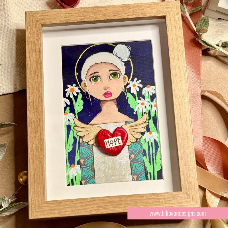 A framed art print featuring a stylized angel illustration with green wings, a white hat, and a red heart that says 'HOPE' on the chest, surrounded by daisies on a dark blue background. The artwork is housed in a light oak wood frame with a white mat, set against a styled background with ribbons and paper.