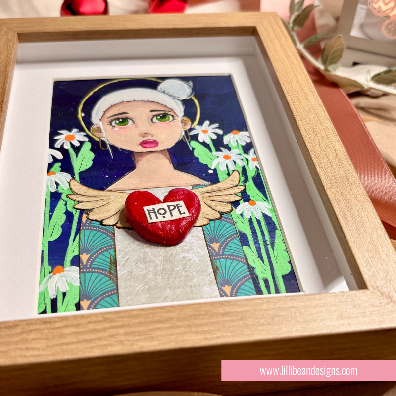 A framed art piece featuring a stylized angel illustration with green wings, white daisies, and a red heart in the center that reads 'HOPE'. The artwork is displayed inside a light wood shadowbox frame with a white mat.