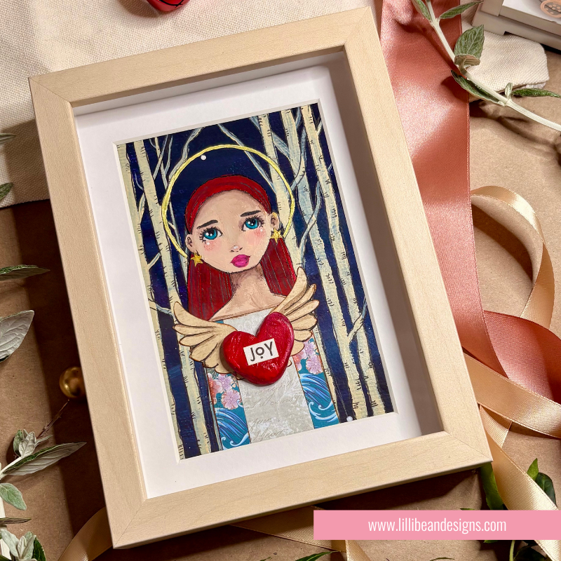 A framed art print featuring a stylized angel with red hair, gold wings, and a red heart that says 'JOY' in white letters, set in a light wood frame on a styled background with ribbons and leaves.