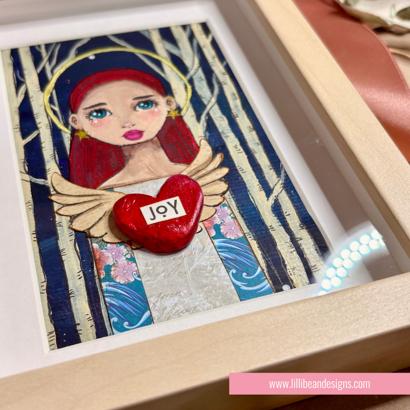 A framed art print featuring a stylized angel with red hair, blue and gold garments, white birch tree silhouettes, and a red heart labeled 'Joy' in the center. The piece is displayed in a light wood frame with a white mat.