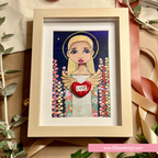 A framed art print featuring a stylized blonde angel with wings, holding a red heart that says 'LOVE' against a dark blue background with colorful dots. The artwork is in a light wood frame and styled on a kraft paper surface with ribbon and greenery.