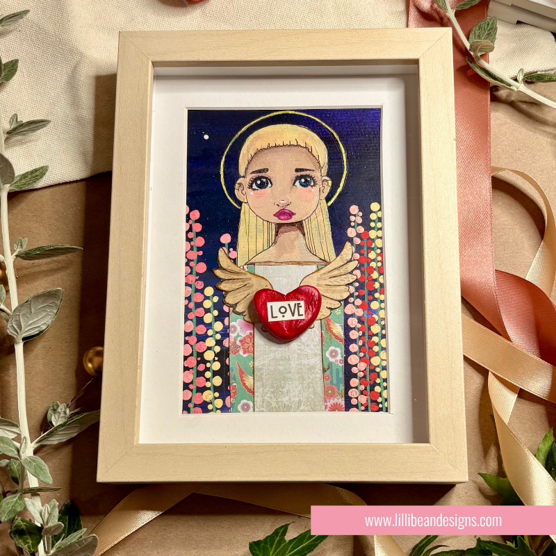 A framed art print featuring a stylized blonde angel with wings, holding a red heart that says 'LOVE' against a dark blue background with colorful dots. The artwork is in a light wood frame and styled on a kraft paper surface with ribbon and greenery.