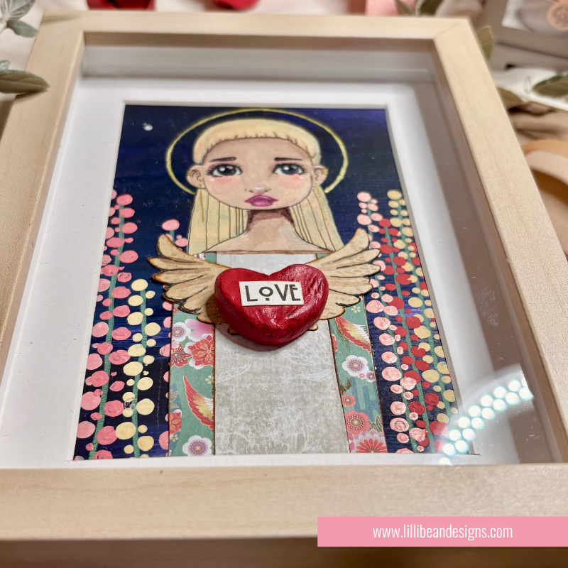 A framed mixed-media art print featuring a stylized angel with blonde hair, wings, and a red heart-shaped stone in the center that reads 'LOVE'. The artwork is housed in a light wood frame with a white mat.