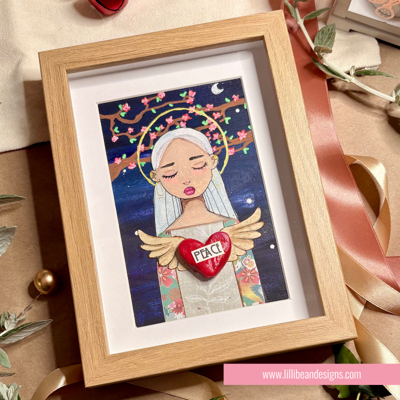 A framed art print featuring a stylized illustration of a woman with white hair, angel wings, and a red heart that says 'PEACE' on a dark blue background, displayed in a light wood frame with decorative ribbons and paper around it.