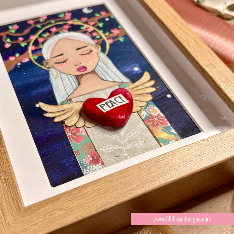 A framed art print featuring a stylized angel with closed eyes, white hair, and wings, holding a red heart labeled 'PEACE' against a dark blue background with floral details. The piece is displayed in a light wood frame with a white mat.