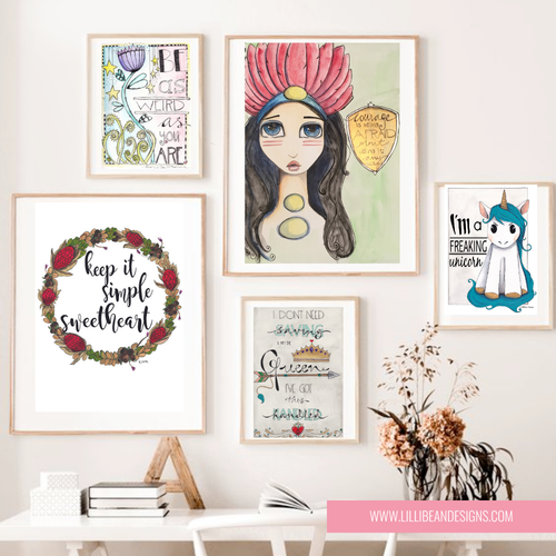 A styled interior wall display featuring five framed art prints with whimsical illustrations and text, including a unicorn, a floral wreath reading 'keep it simple sweetheart,' a pink-haired woman, and decorative lettering. A white table, books, and dried flowers sit below against a light wall.