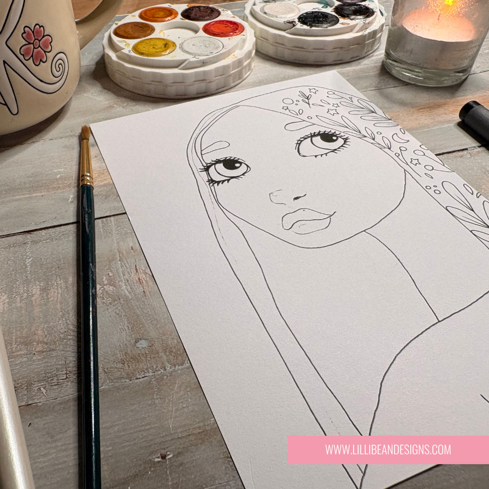A black line art portrait illustration of a woman's face with floral hair details on a white sheet of paper, styled on a wooden table with a paintbrush, paint palettes, and a lit candle.