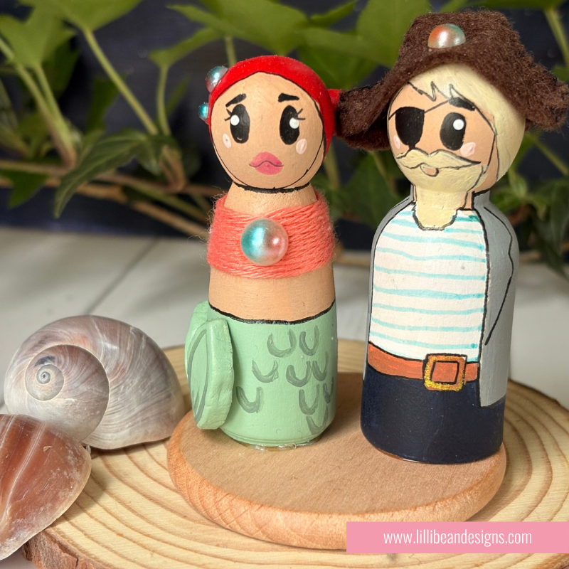 Handmade wooden mermaid and pirate doll couple figurines on a round wooden base, styled with seashells and green leaves in the background.