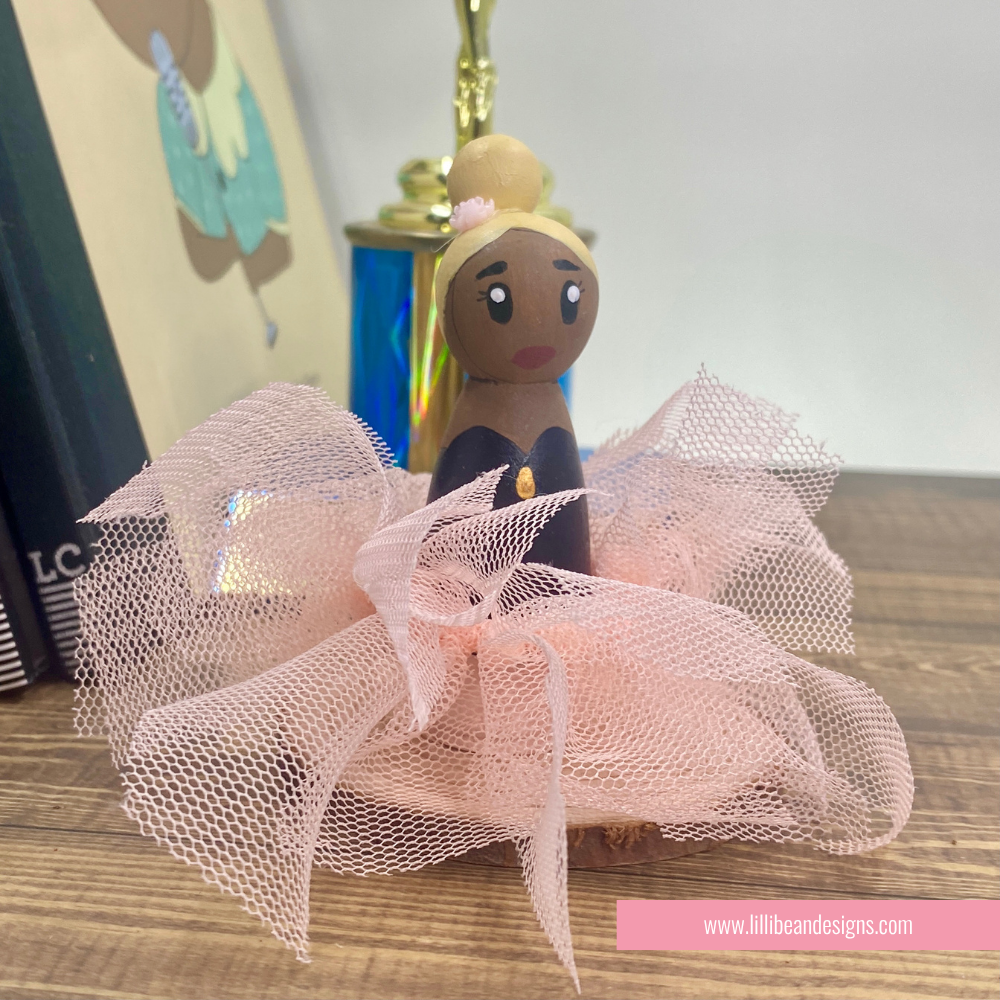 A hand-painted wooden doll figurine with a blonde bun hairstyle, dark brown skin, and a fluffy pink tulle tutu, displayed on a wooden surface.