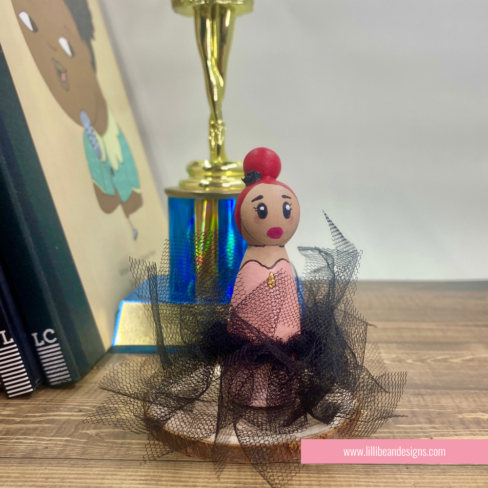 A hand-painted wooden peg doll styled as a ballerina, featuring a pink dress, black tulle skirt, and brown hair in a bun, displayed on a wooden surface.