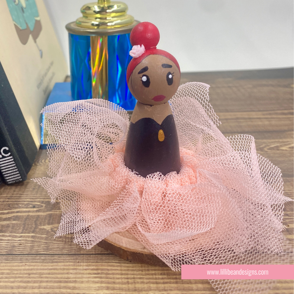 A handcrafted wooden doll figurine with a brown face and hands, red hair, and a black dress, styled with a pink tulle tutu skirt on a wooden surface.