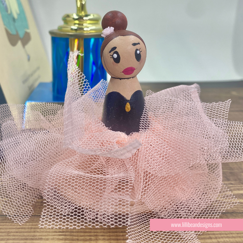 A hand-painted wooden peg doll styled as a ballerina, featuring a pink dress, black tulle skirt, and brown hair in a bun, displayed on a wooden surface.