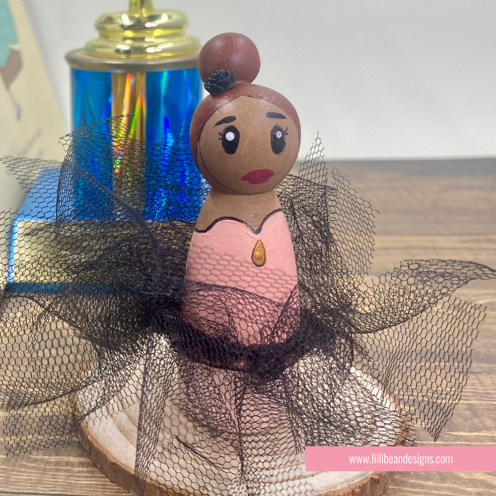 A hand-painted wooden peg doll styled as a ballerina, featuring a pink dress, black tulle skirt, and brown hair in a bun, displayed on a wooden surface.