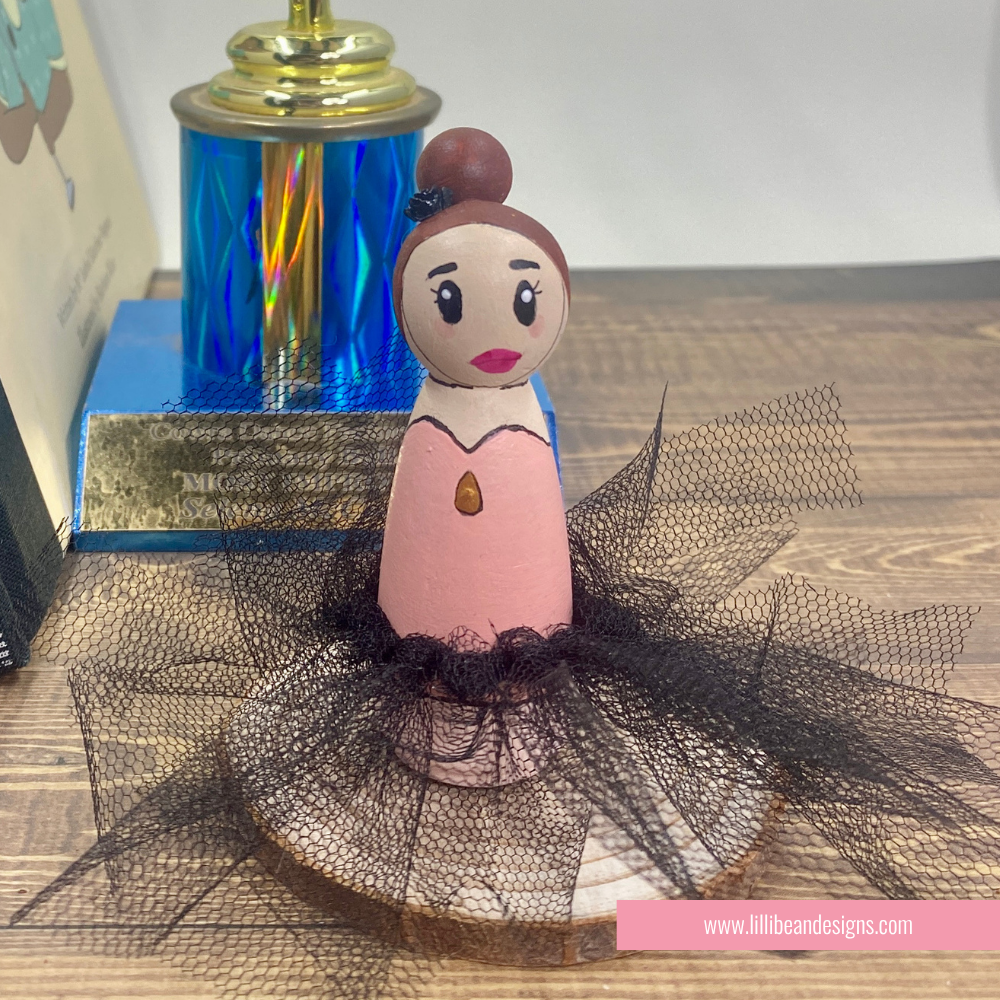 A handcrafted wooden ballerina doll with a pink dress, black tulle tutu, and brown hair bun, displayed on a wooden surface with a trophy in the background.