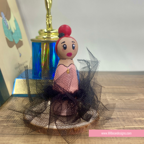 A handcrafted wooden doll figurine styled as a ballerina, featuring a pink dress, black tulle tutu, and a red hair bun, displayed on a wooden surface with a trophy in the background.