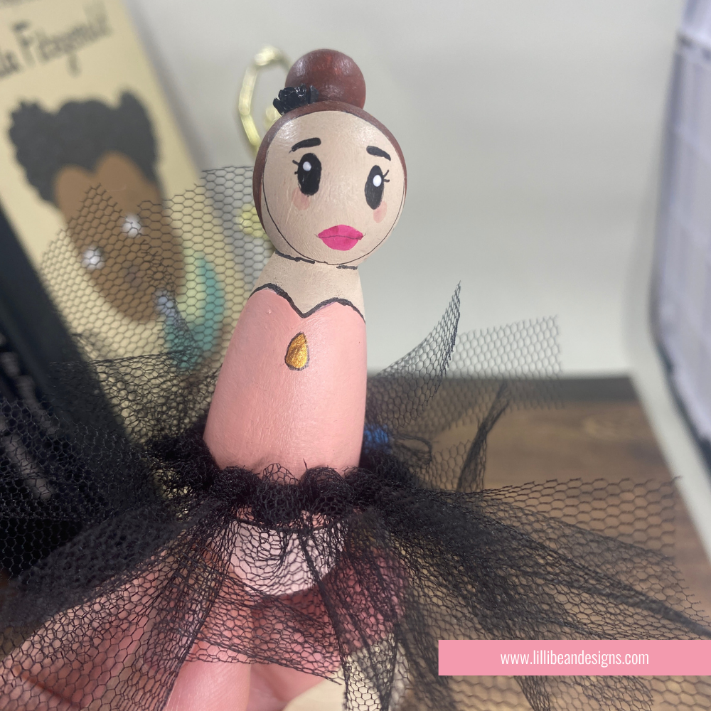 A handcrafted wooden ballerina figurine with a painted face, brown hair bun, pink dress, and black tulle skirt, shown close-up with a finger supporting it.