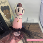 A handcrafted wooden ballerina figurine with a painted face, brown hair bun, pink dress, and black tulle skirt, shown close-up with a finger supporting it.
