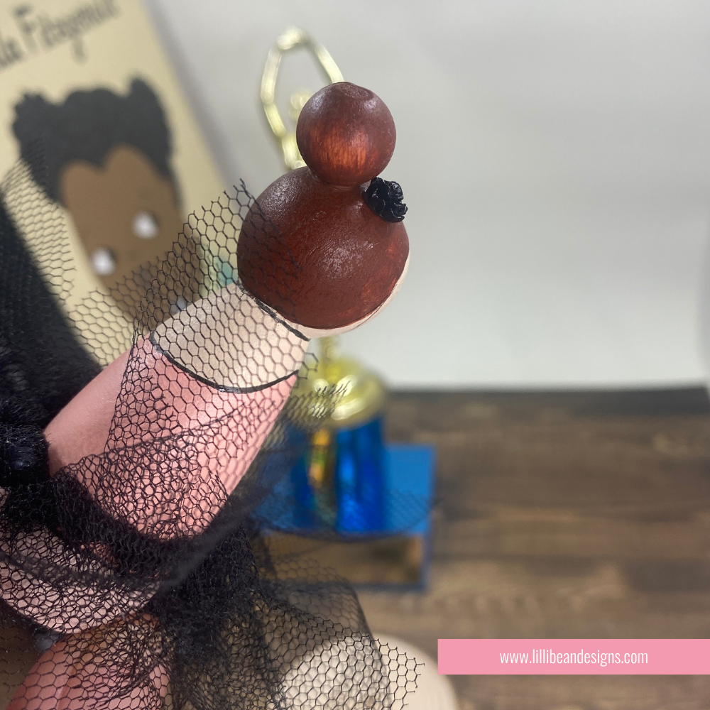 A close-up of a doll or figurine with a dark brown wooden head, black tulle dress, and gold ring, held in front of a blurred background with a book and small blue object.