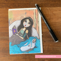 GREETING CARD Illustration of a woman with a unicorn and a book on a wooden surface with a pen.