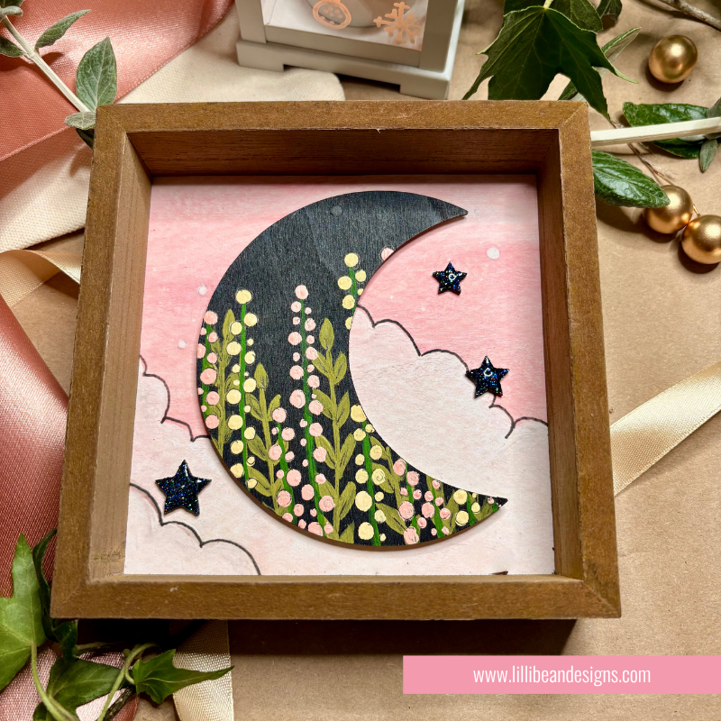 A square wooden shadow box frame displaying a handmade paper craft of a crescent moon filled with green leaves and pink flowers, set against a pink sky with clouds and small star details, styled with ribbon and greenery.
