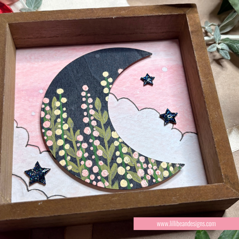 A decorative wooden shadow box featuring a crescent moon and stars. The crescent moon is decorated with green leaves and pink and yellow flowers, set against a pink sky with white clouds.