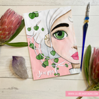 Journal with a face design and greenery on a wooden surface with flowers and a pen.