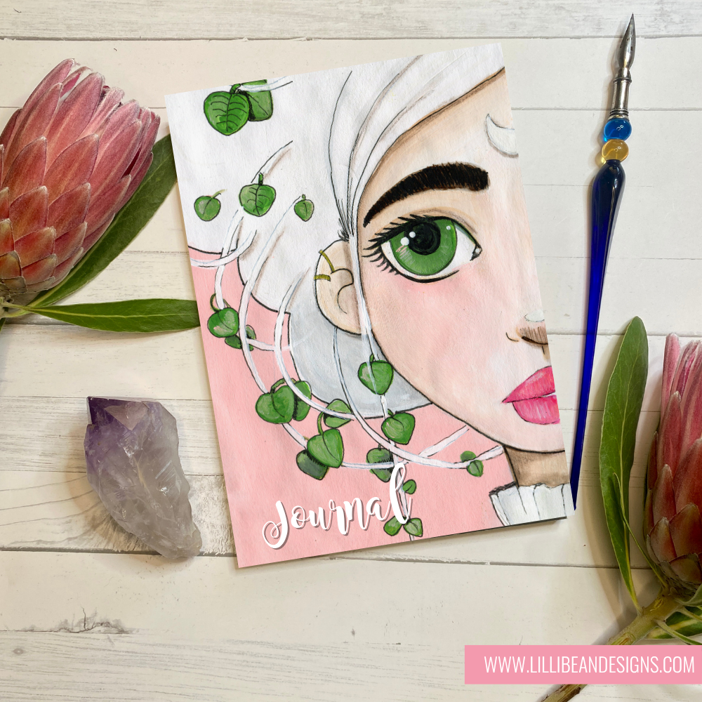 Journal with a face design and greenery on a wooden surface with flowers and a pen.