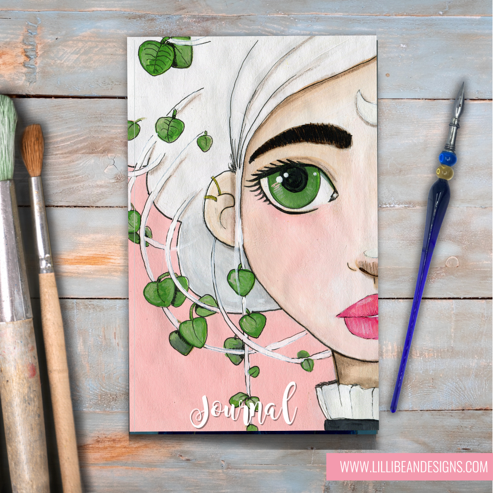 Journal cover with artistic face design and greenery on a wooden surface with painting tools.