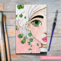 Journal cover with artistic face design and greenery on a wooden surface with painting tools.