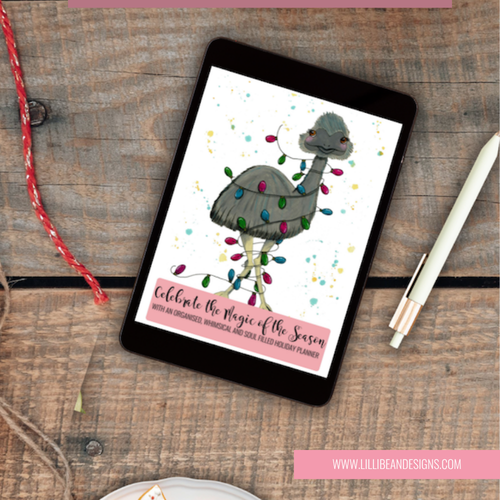 A tablet on a rustic wooden tabletop displaying a holiday planner cover with a gray Christmas tree decorated with colorful lights. A white pen rests to the right, and pink branding text at the bottom reads www.