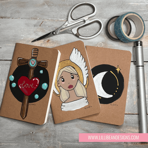 Three decorated notebooks with artistic designs on a wooden surface with scissors and tape.