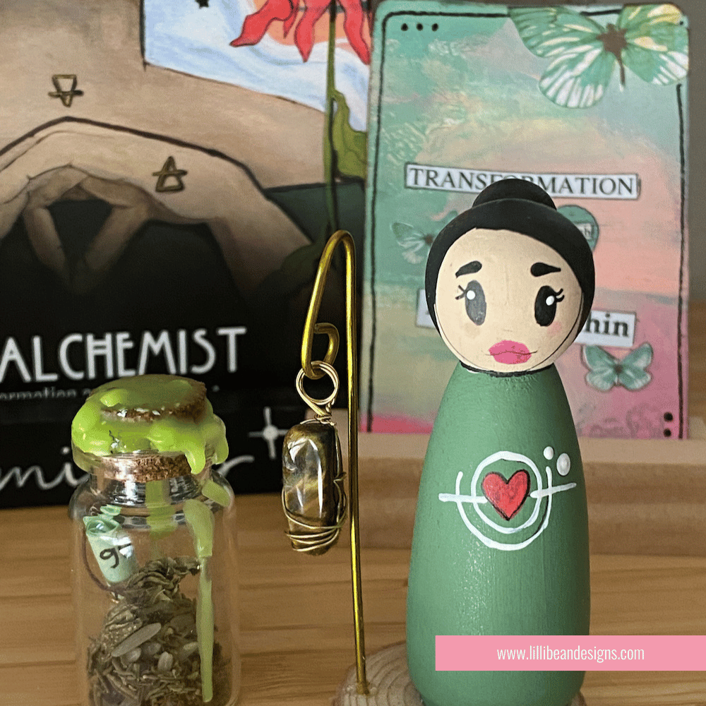 Green peg doll with a heart design, small bottle with green substance, and gold pendant on a wooden surface.