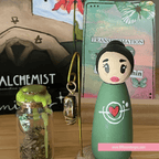 Green peg doll with a heart design, small bottle with green substance, and gold pendant on a wooden surface.