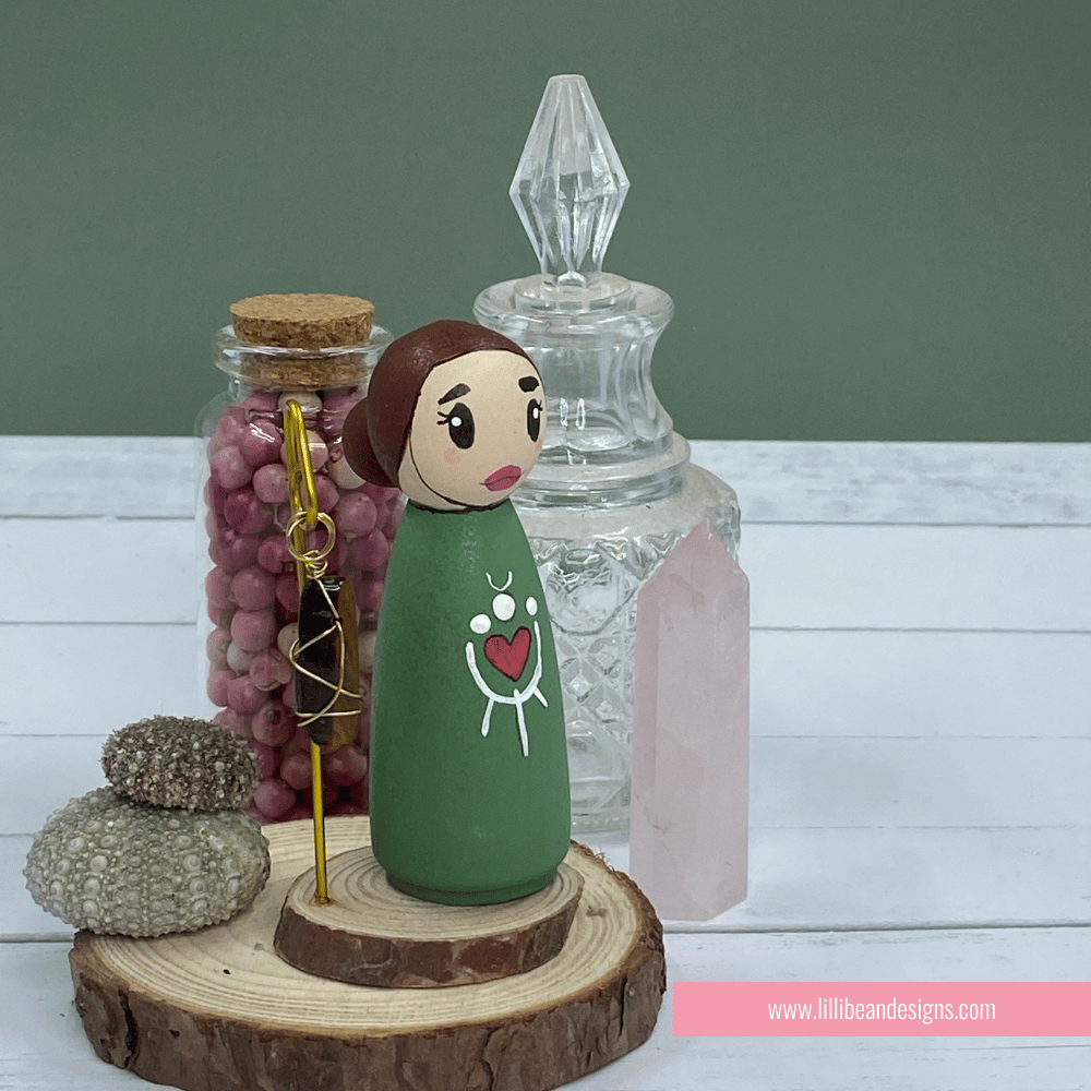 Peg doll with green dress and heart design on a wooden stand with decorative jars and stones.