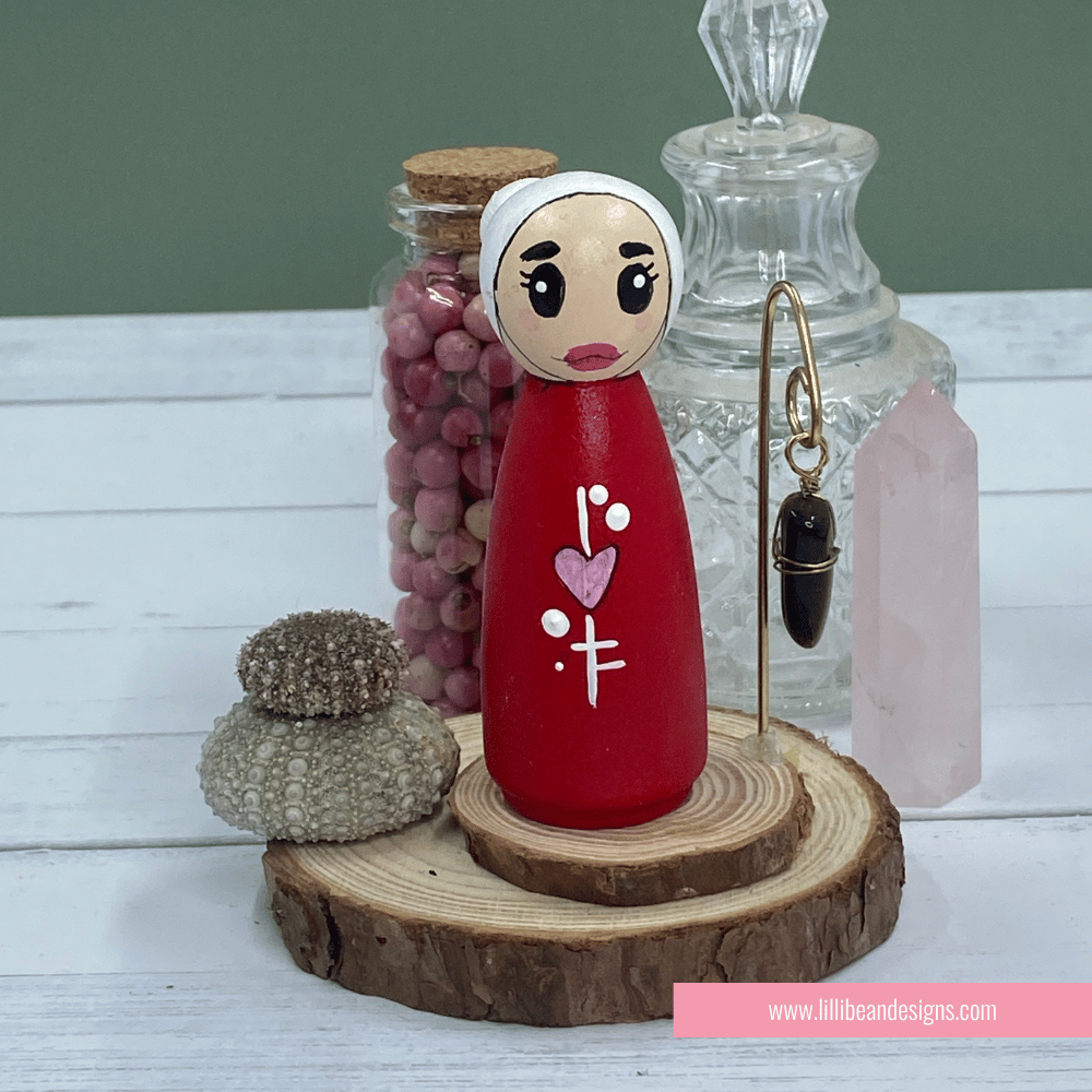 Wooden peg doll with red body and white face on a wooden stand, surrounded by decorative items.