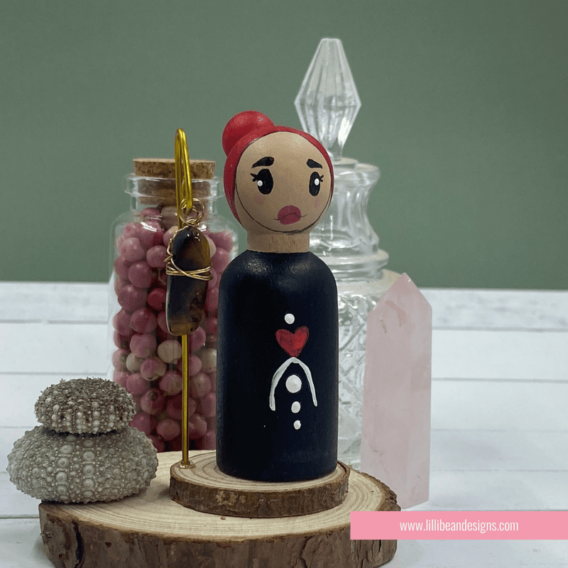 Peg doll with red hair and black body on a wooden stand with decorative items in the background.
