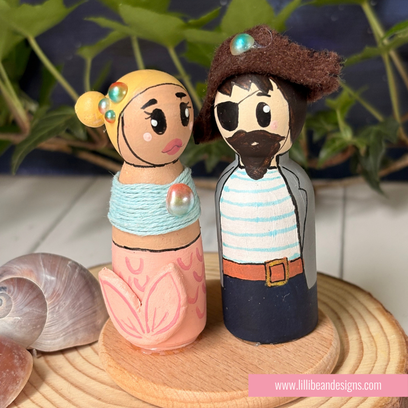 Hand-painted wooden peg dolls styled as a mermaid and a pirate, displayed on a wooden coaster with seashells and green foliage in the background.