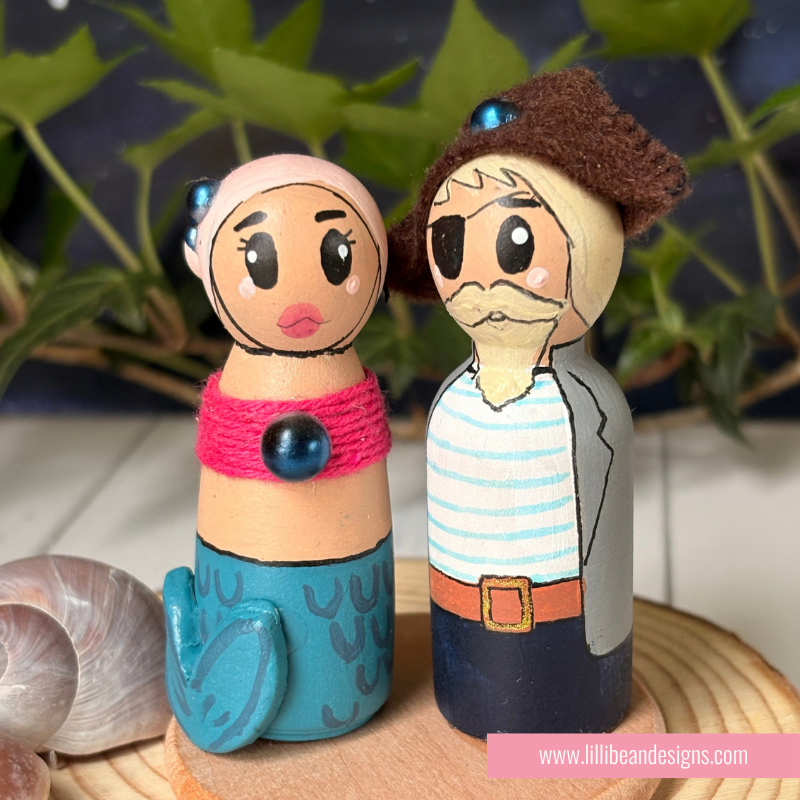 Hand-painted wooden peg dolls representing a couple, one with a mermaid tail and pink top, and the other with a brown hat and striped shirt, displayed on a wooden slice with a shell in the foreground.