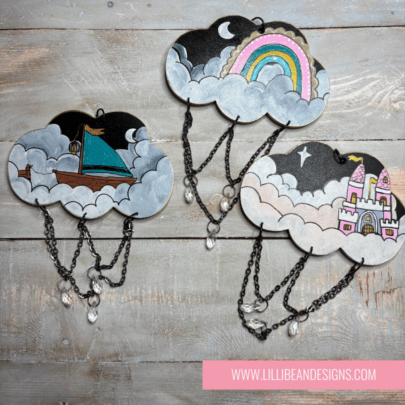 Decorative cloud-shaped hooks with illustrations on a wooden surface, labeled with rainbow image, boat and castle. LilliBean Designs