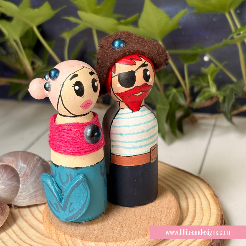 Handcrafted wooden peg dolls styled as a mermaid and pirate couple, standing on a round wooden slice with seashells and green leaves in the background.