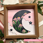 A square wooden shadow box containing a whimsical handmade paper art design of a crescent moon with pink flowers, green stems, and black stars on a pink background, surrounded by decorative leaves and ribbons.