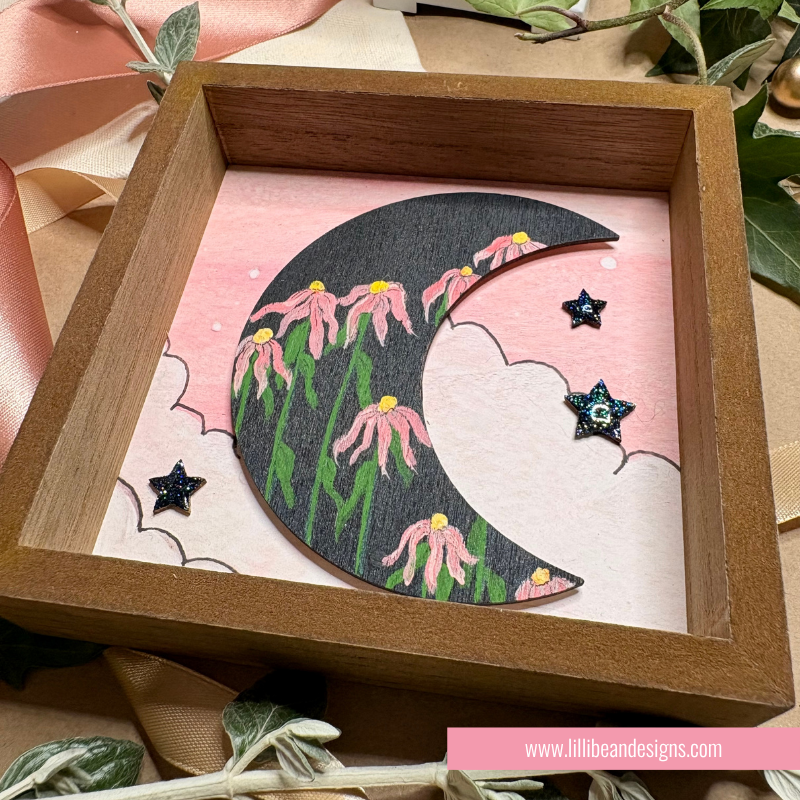 A square wooden shadow box display featuring a black crescent moon decorated with pink flowers and green leaves, set against a pink cloud-patterned backing. The box is styled with decorative ribbons and leaves.
