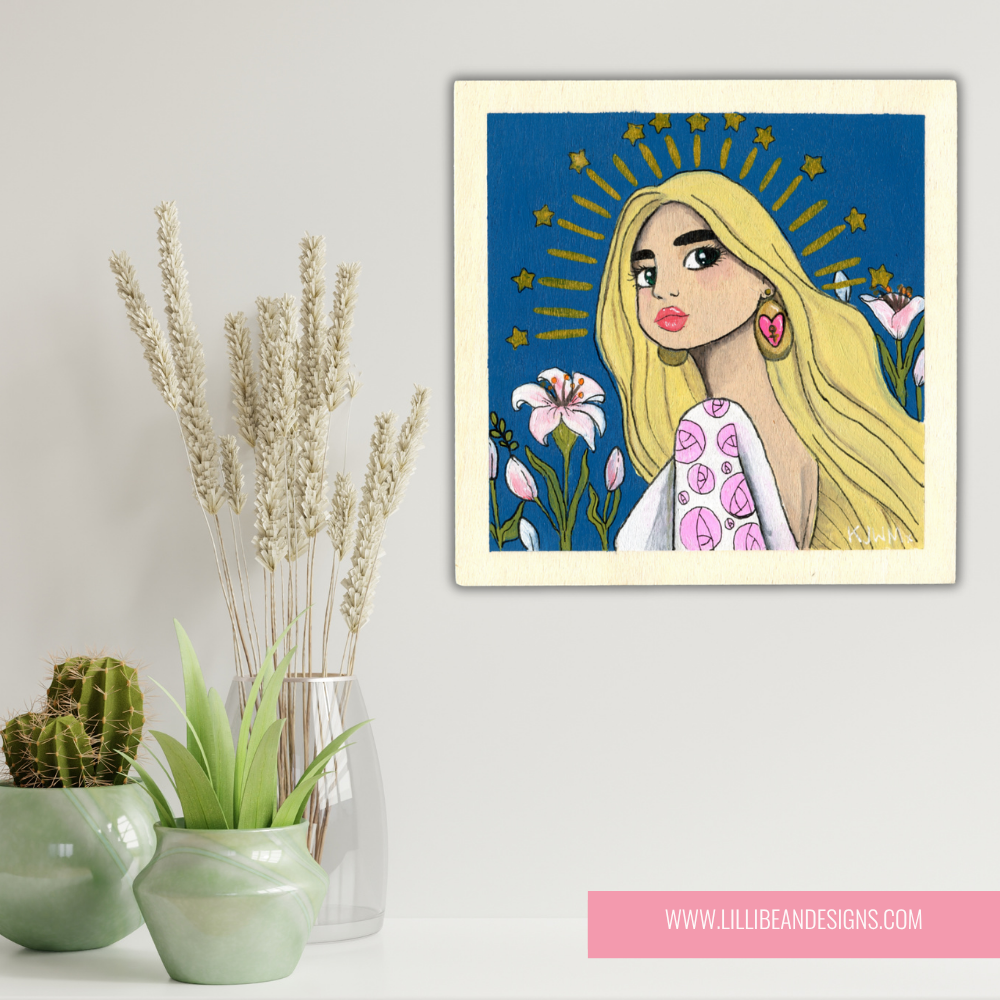 Framed artwork of a woman with blonde hair and floral design, placed on a white surface with decorative plants.