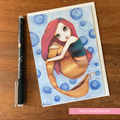 Illustration of a mermaid on a card with a pen next to it on a wooden surface.