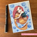 Illustration of a mermaid on a card with a pen next to it on a wooden surface.