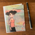 GREETING CARD with an illustration of a girl on a wooden surface with a pen next to it